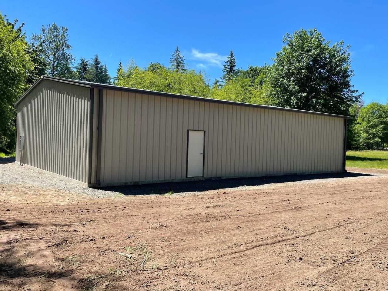 Beige metal storage building with a white door, set on a gravel lot surrounded by trees and blue sky