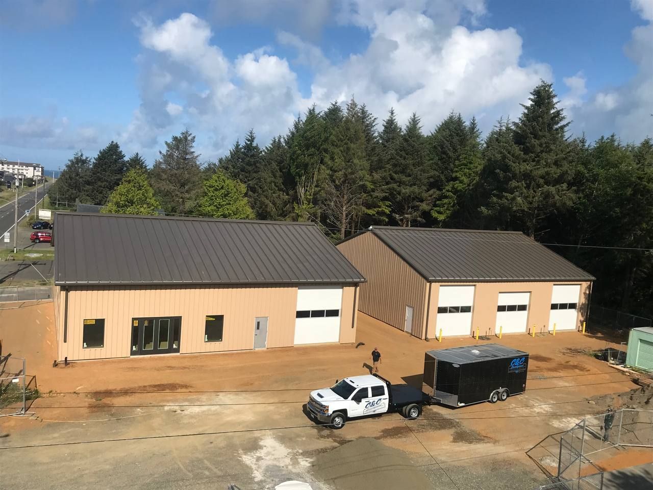 Two tan buildings with dark roofs, a white truck and blue trailer in a dirt lot surrounded by trees.