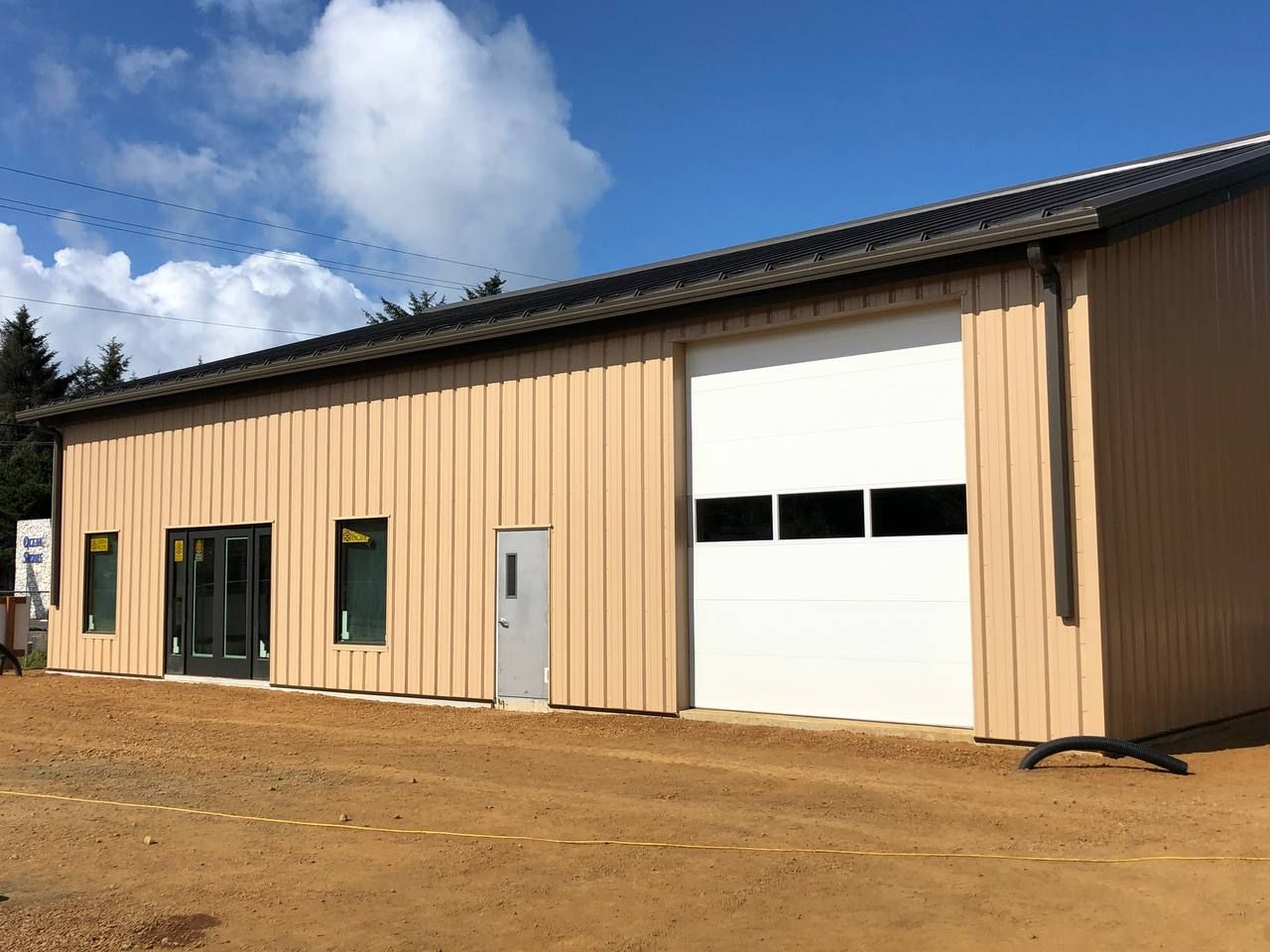 Tan metal building with white garage door and small windows on a dirt lot under a blue sky