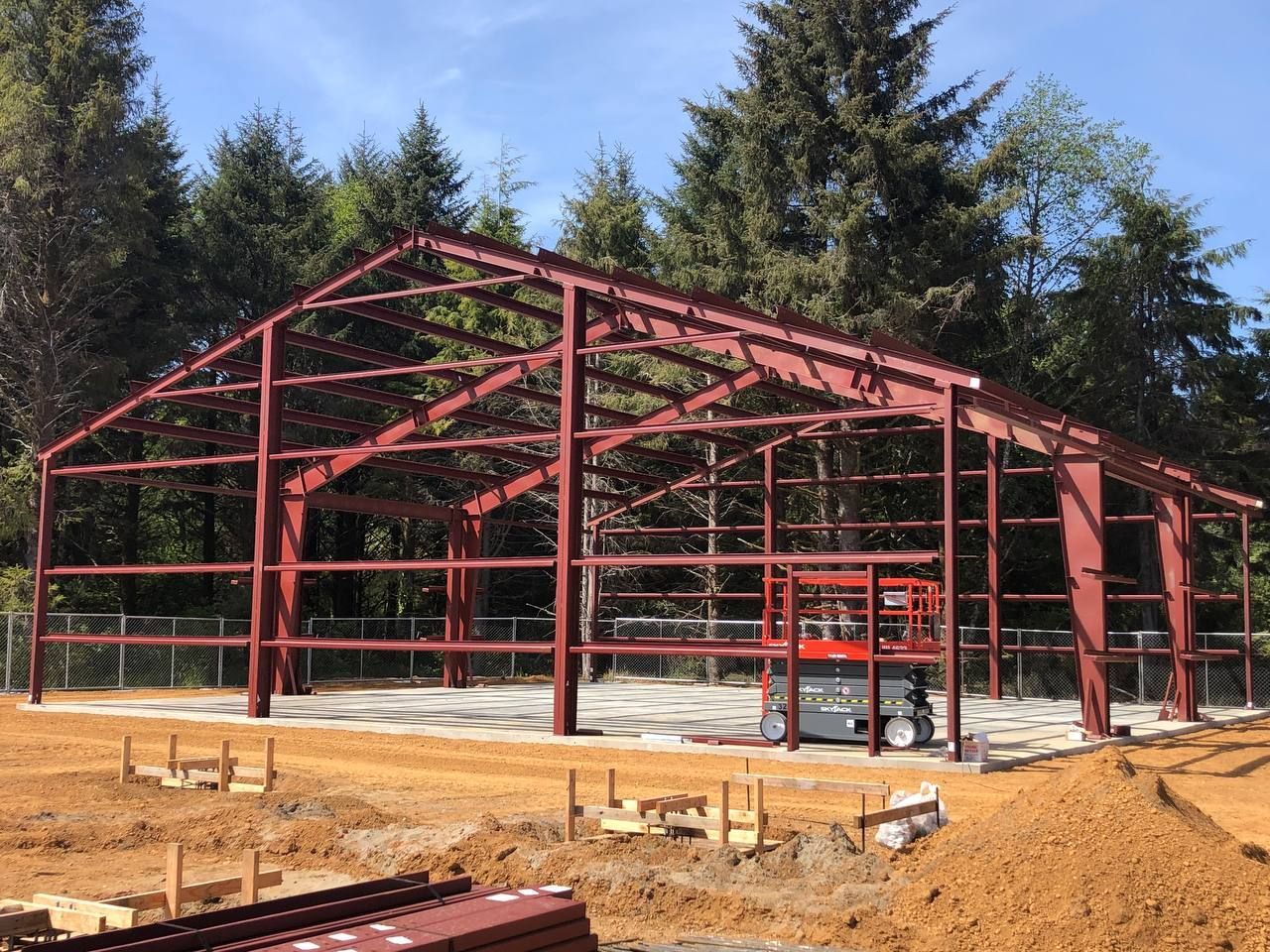 Red steel-framed building under construction on a concrete slab with dirt foreground and trees behind.