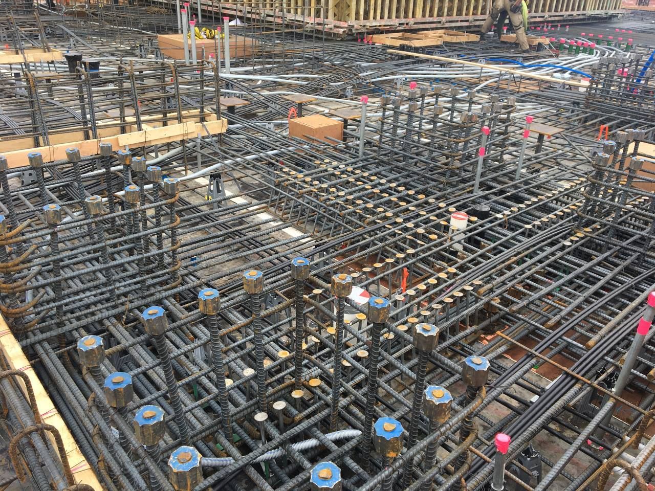 Rebar grid and formwork at a construction site, with concrete footings and scaffolding in the background