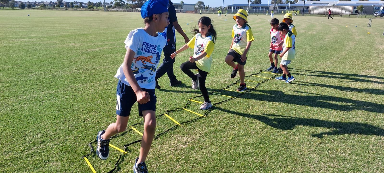 Children practice footwork drills on an agility ladder set up on a grassy field.