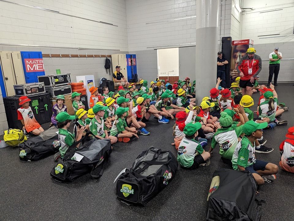 A large group of children in green and white sports uniforms sits on the floor in a locker room with sports bags.