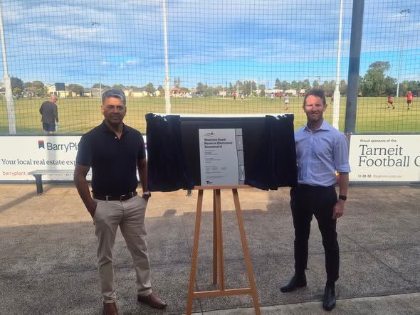 Two men stand on either side of a plaque on an easel at the Tarneit Football Club, during a facility opening ceremony.
