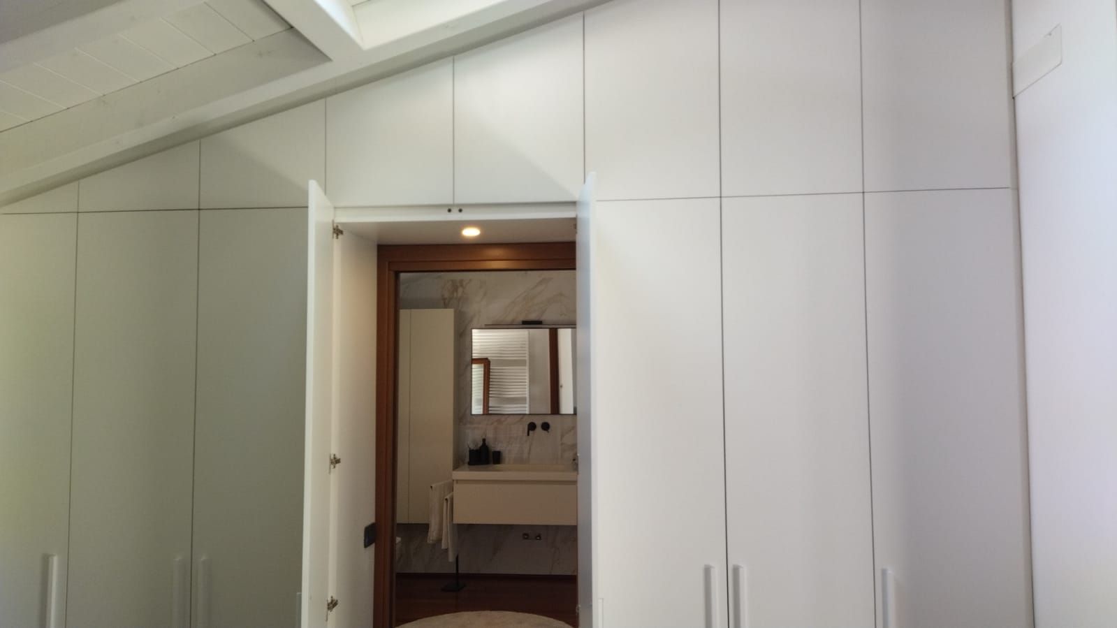 arredo armadio su misura, con moduli contenitivi che seguono le linee del tetto. ante centrali passanti per accesso alla zona bagno.  illuminazione led interno passaggio. Arredo bagno Cerasa sospeso in laccato opaco e lavabo in marmo resina. realizzazione in contesto villa a Como.