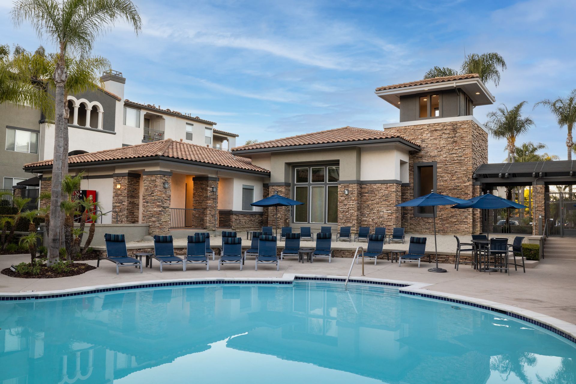 Outdoor apartment pool area with blue lounge chairs and umbrellas in front of stone-clad buildings at Alize Apartment Homes in Aliso Viejo, CA.