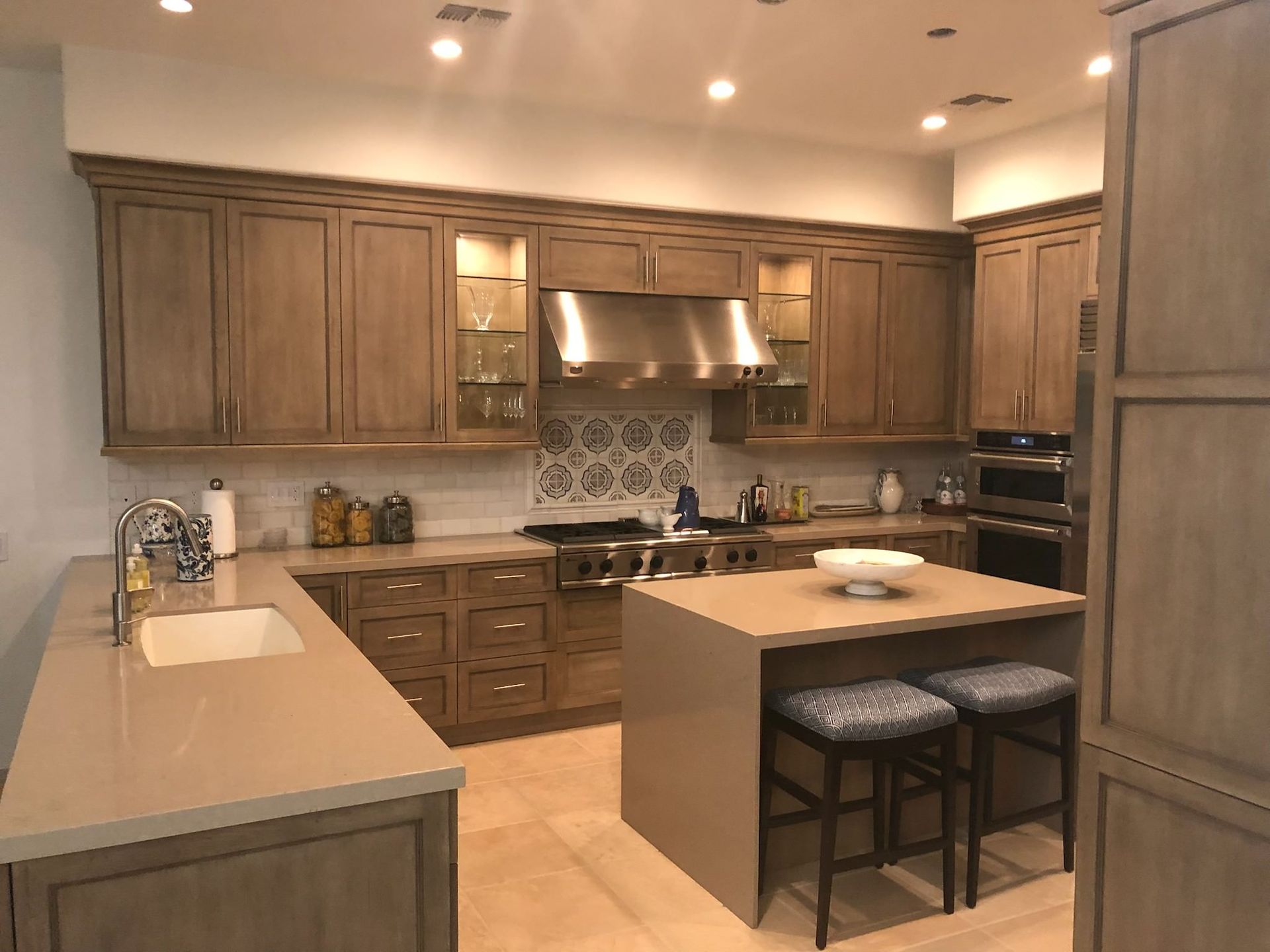 Kitchen with light wood cabinets, gray countertops, stainless steel range hood, and island with stools.