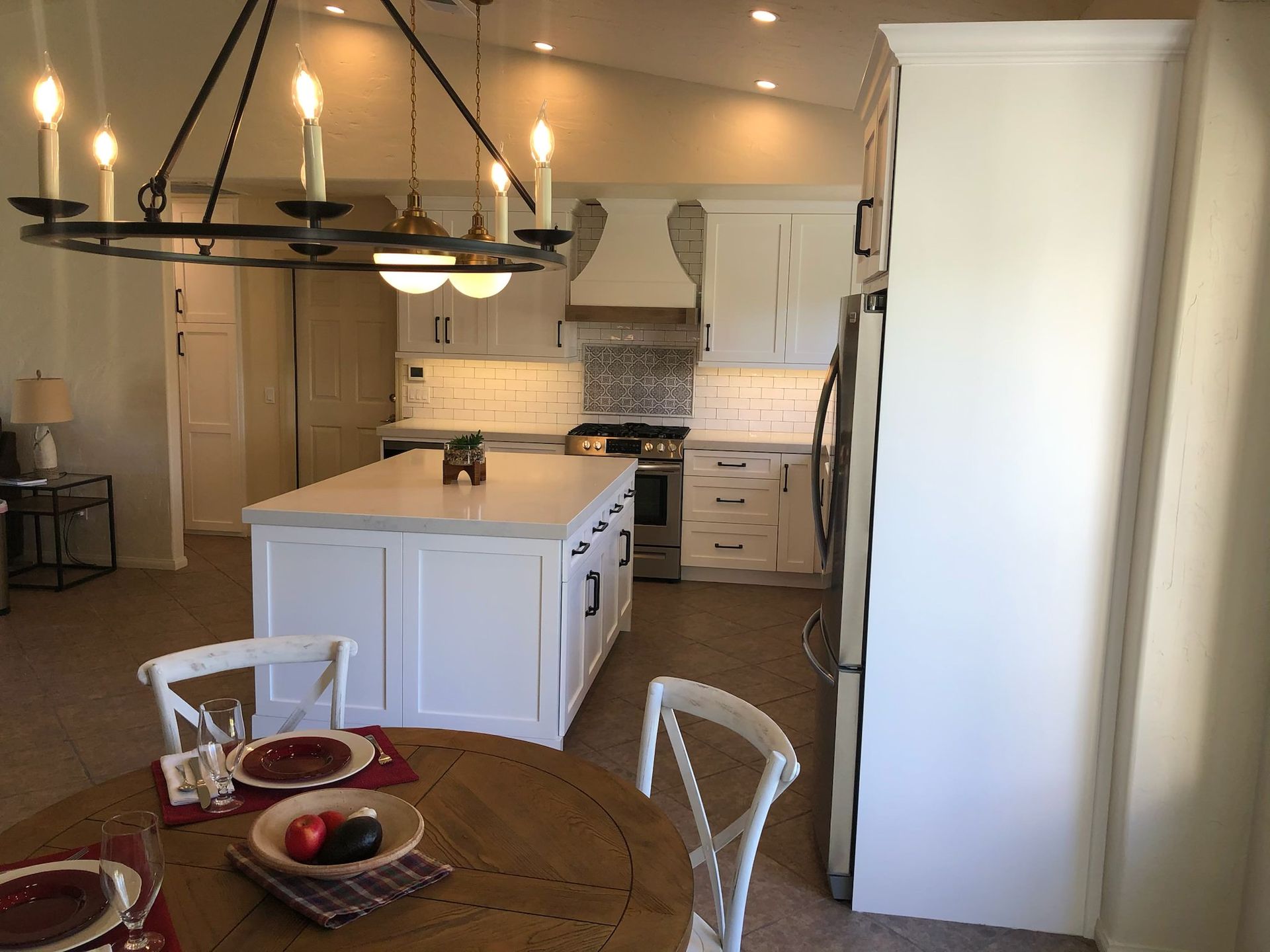 A kitchen with white cabinets, island, and appliances. A dining table and chandelier are in the foreground.