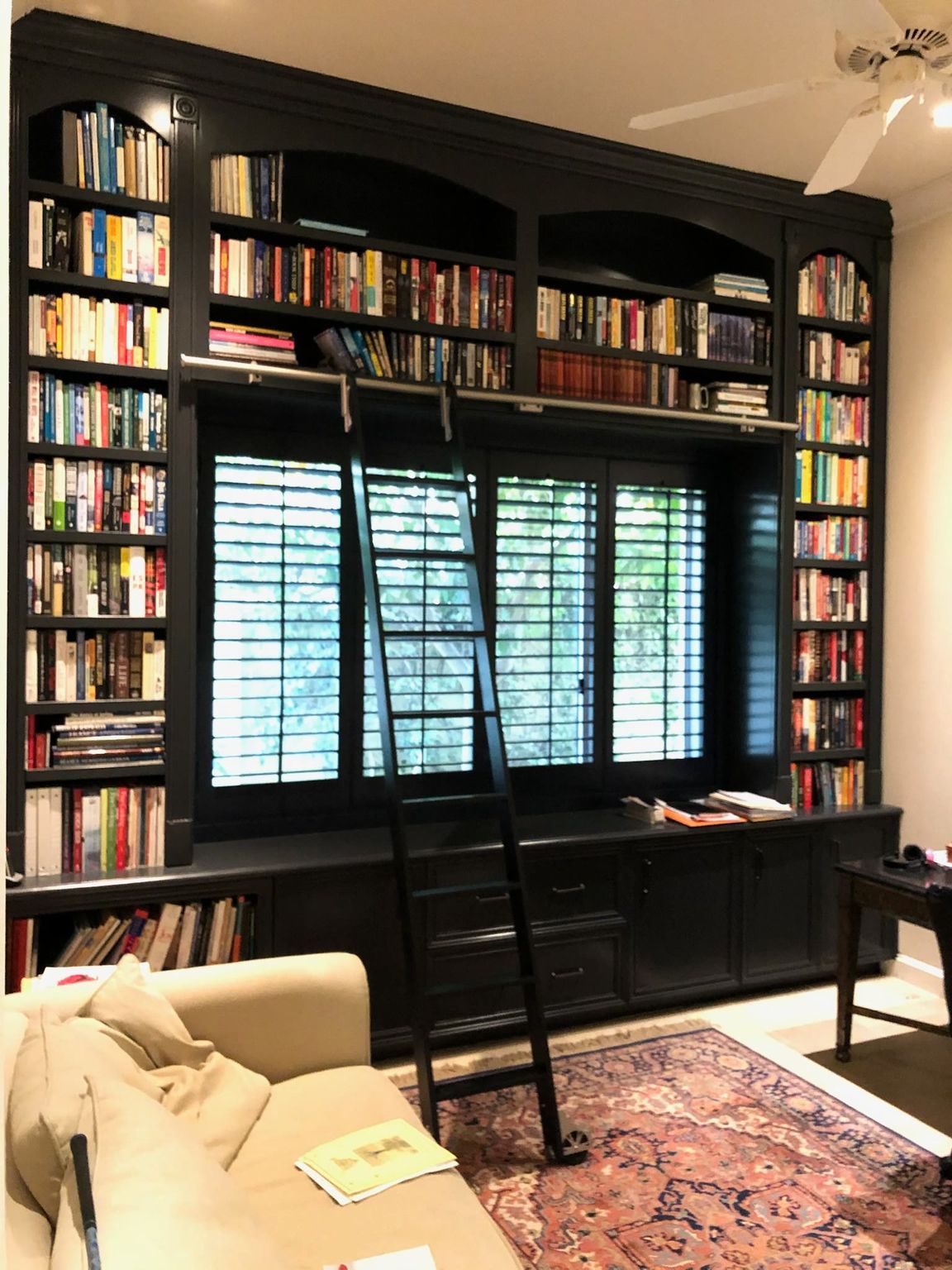 Black bookshelf filled with books, a rolling ladder, and a window with closed shutters. Beige couch and rug in front.