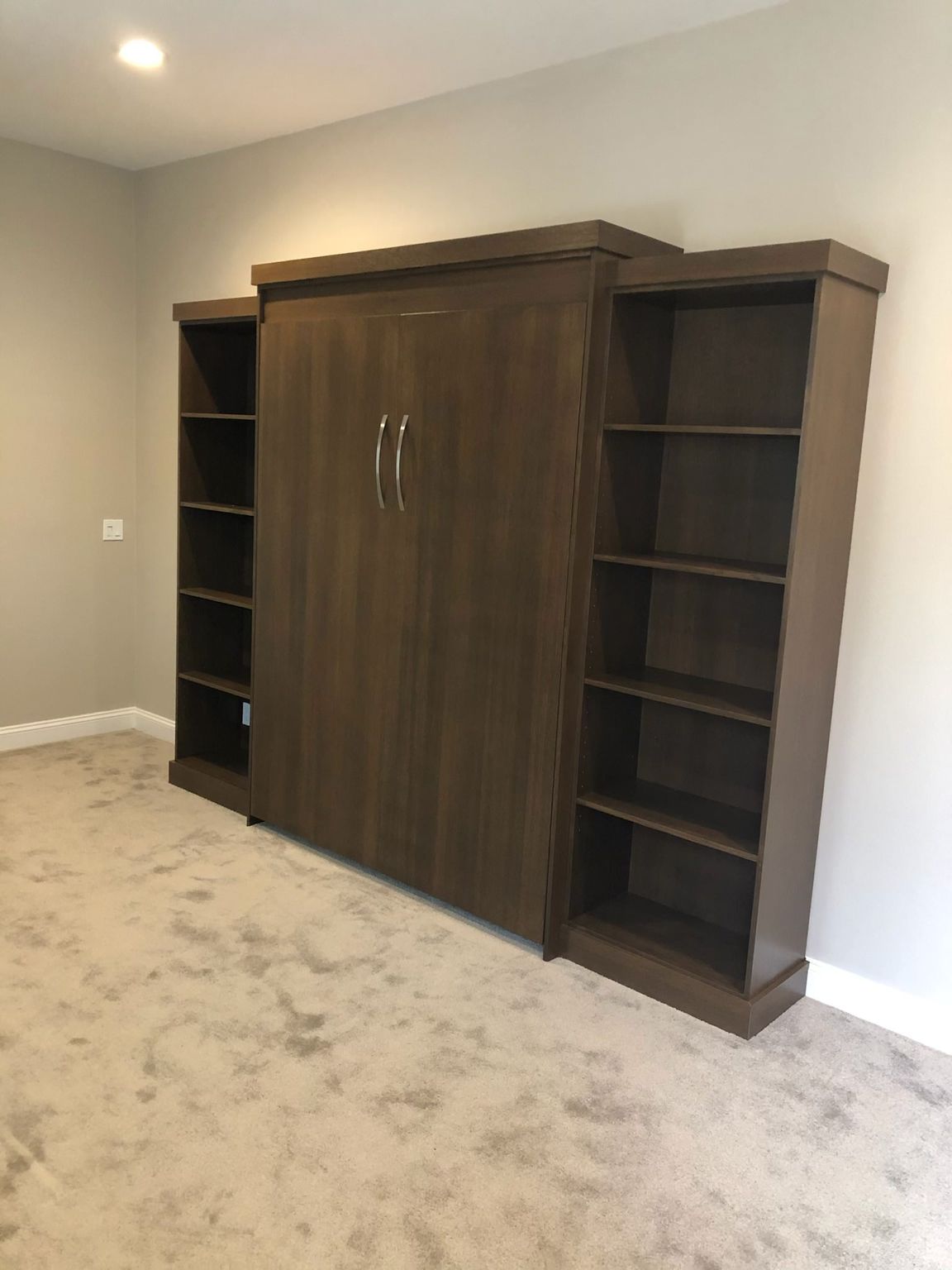 Wall bed with bookshelves on either side, dark wood, closed. Set against a light gray wall on a carpeted floor.