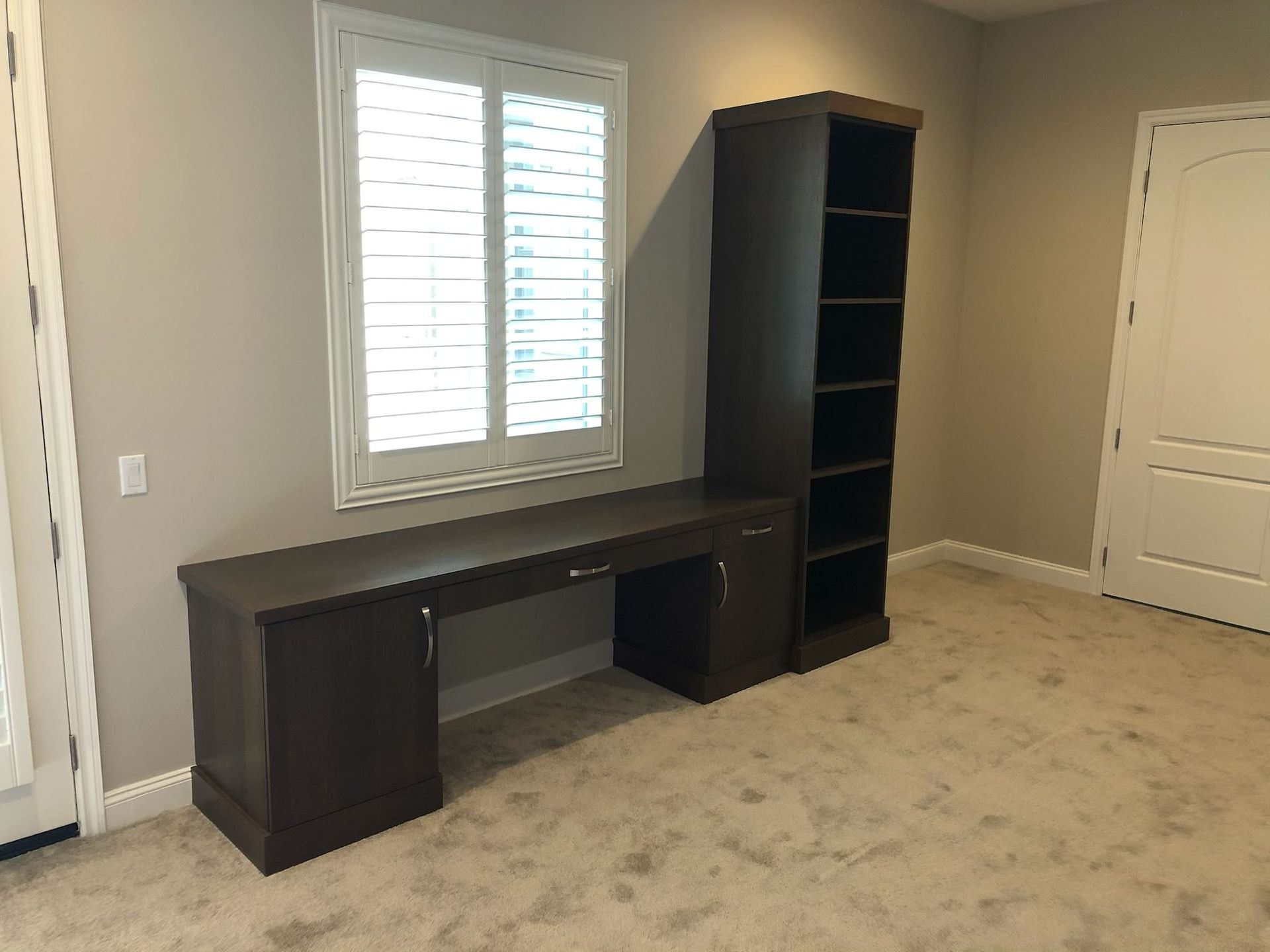 Dark brown desk and bookshelf unit against a beige wall, with a shuttered window and closed door.