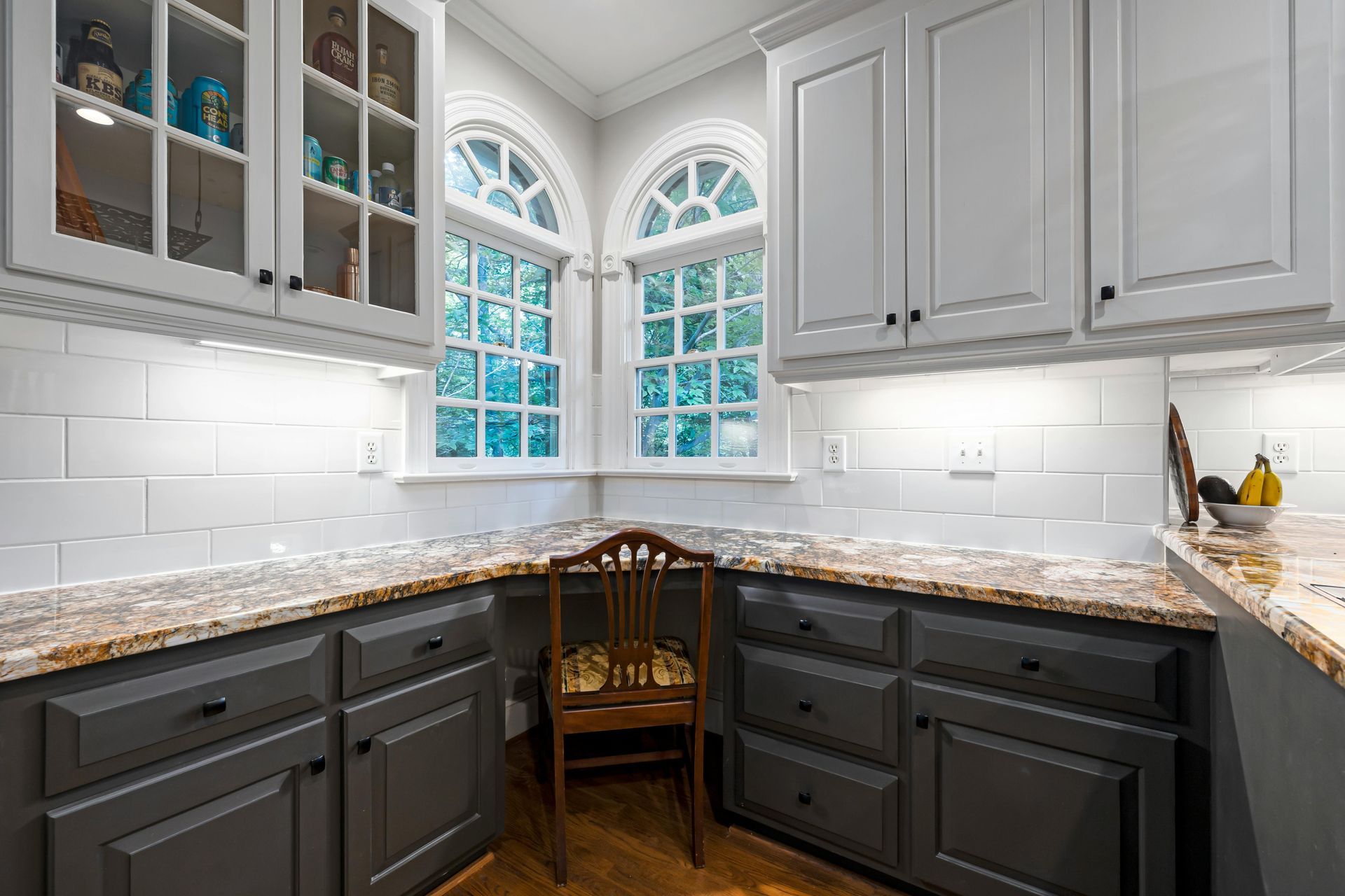 Gray and white kitchen with arched windows, countertops, cabinets, and a chair.
