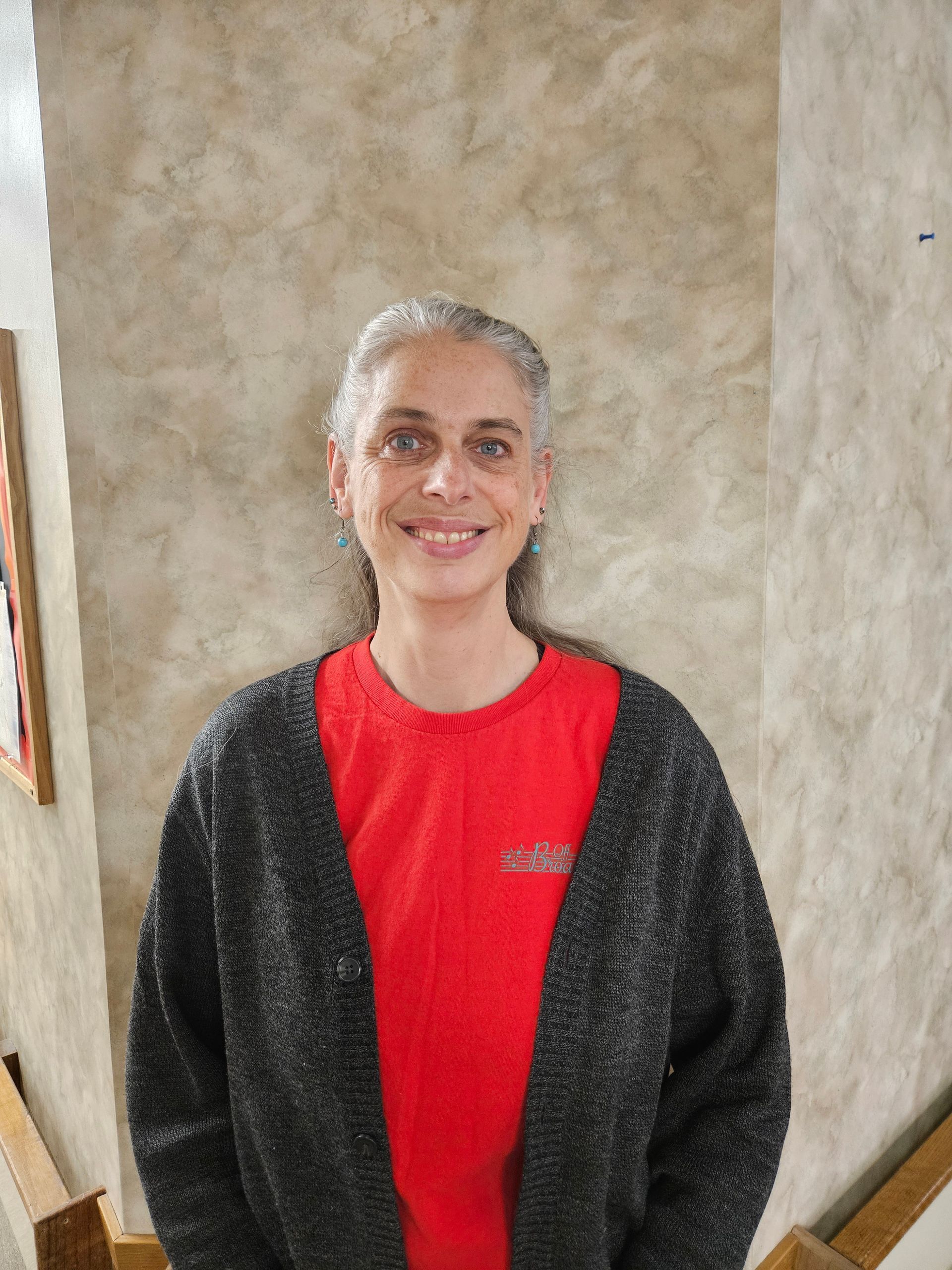 Woman with silver hair smiles in front of a textured beige wall, wearing a red shirt and gray cardigan.