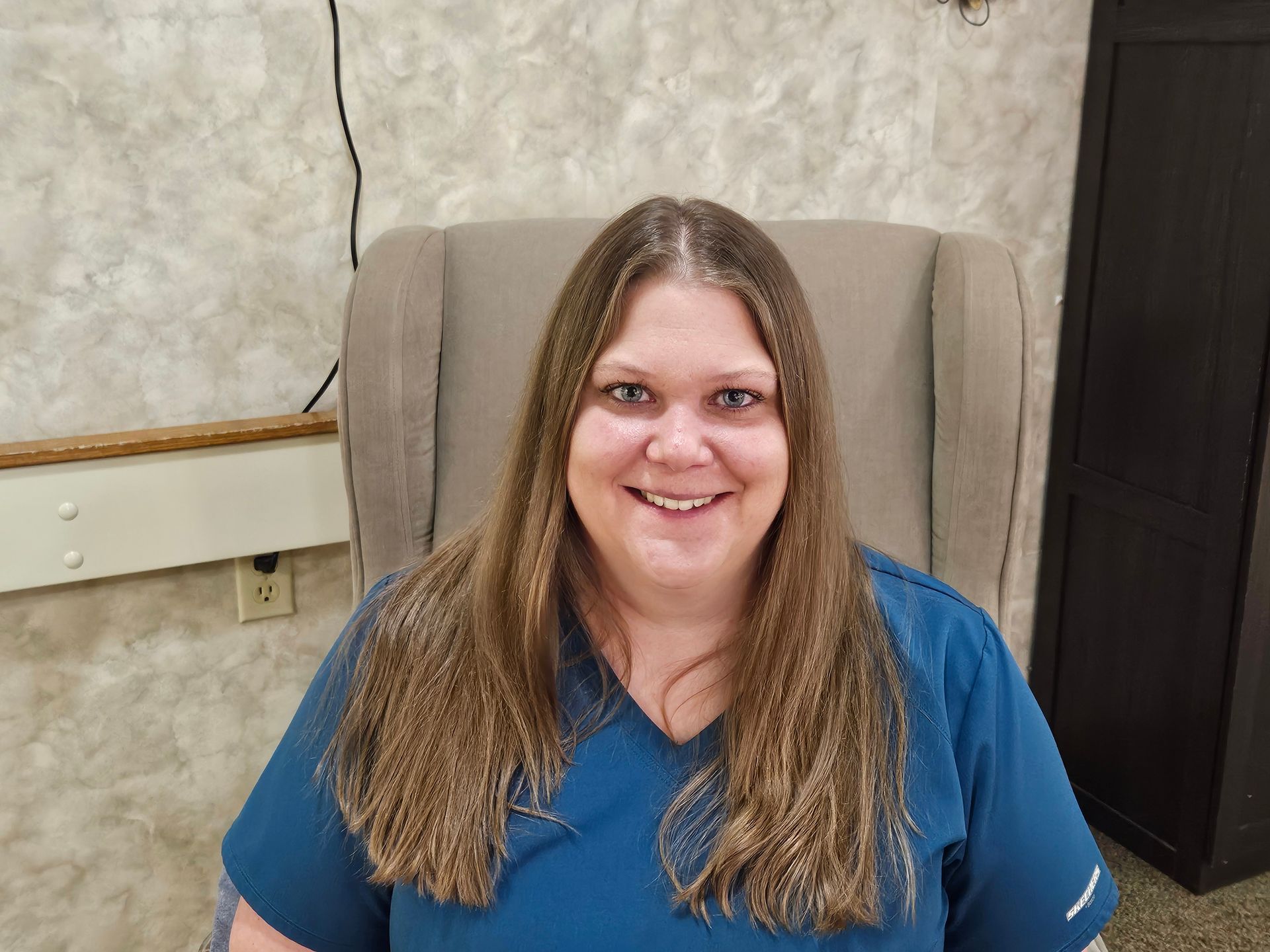 A woman in a blue scrub top is sitting in a chair and smiling.