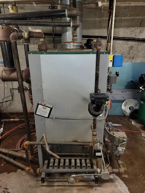 A gas-fired boiler in a basement setting. Gray metal unit with black pipes, valves, and yellow electrical box.