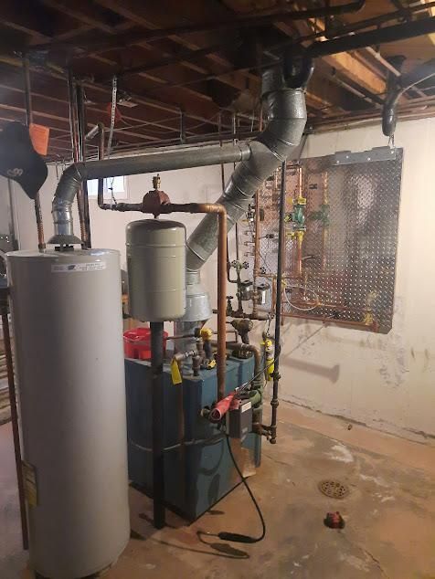 A boiler and water heater system in a basement with copper pipes and a metal vent, on a concrete floor.