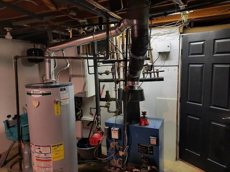 Basement mechanical room with a water heater, furnace, and black door. Pipes and ductwork are visible.