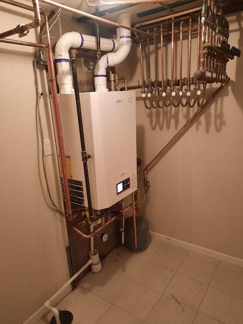 White wall-mounted boiler with copper piping, white ventilation pipes, and a copper manifold in a basement corner.
