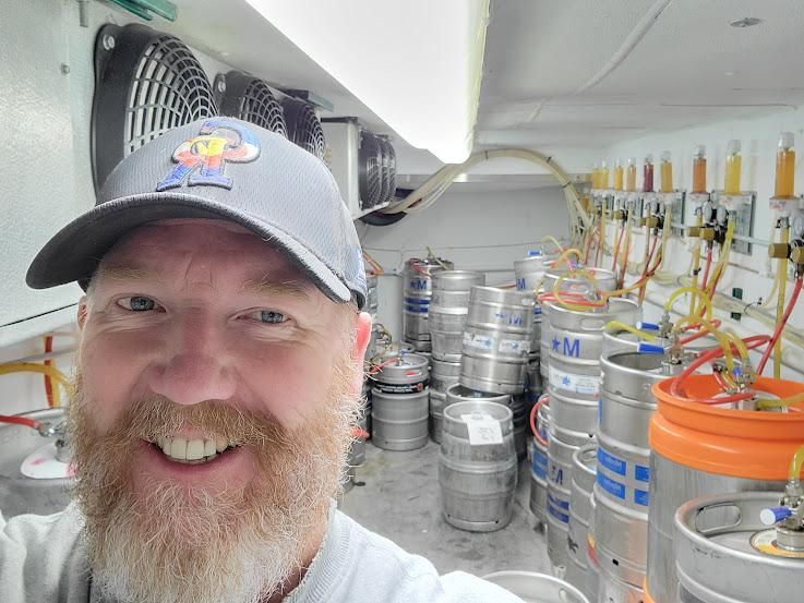 Man with beard smiles in a beer keg cooler; kegs and tap lines fill the background.