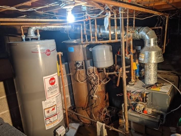 Water heaters and furnace in a basement, with various pipes and ductwork.