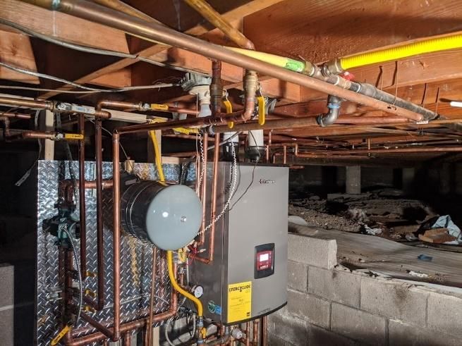 Plumbing and heating system in crawlspace, copper pipes, a gray tank, and yellow gas lines against a wood ceiling.