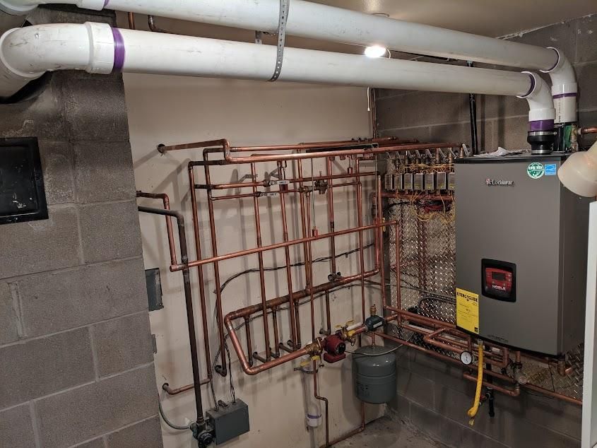 Basement with boiler, copper pipes, and white PVC pipes. Concrete block walls.