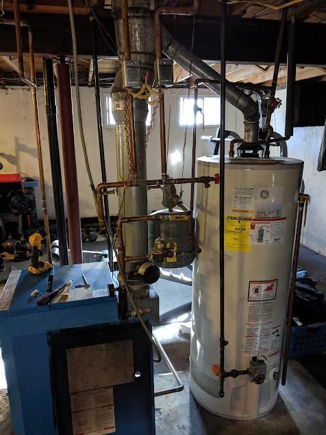 Boiler and water heater in a basement. White water heater, blue boiler, copper pipes, and a light-filled setting.