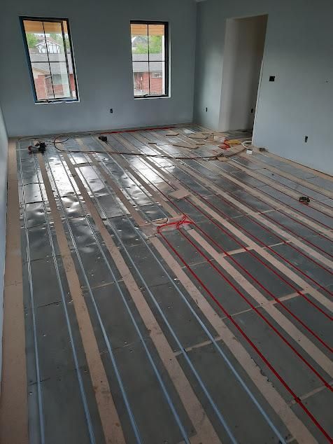 Installation of radiant floor heating in a room with gray walls, two windows, and red heating wires.