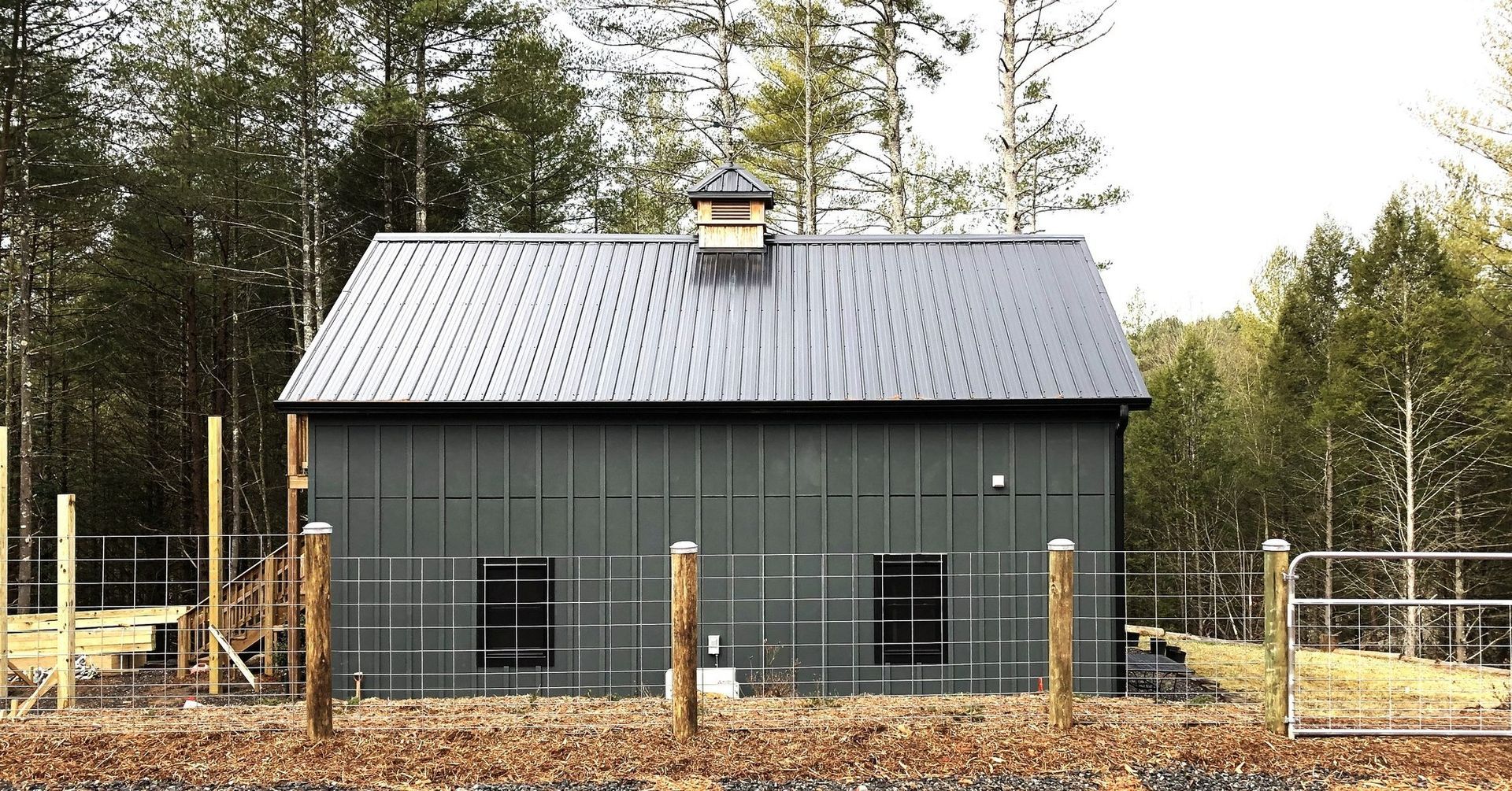 A black barn is surrounded by a fence and trees.