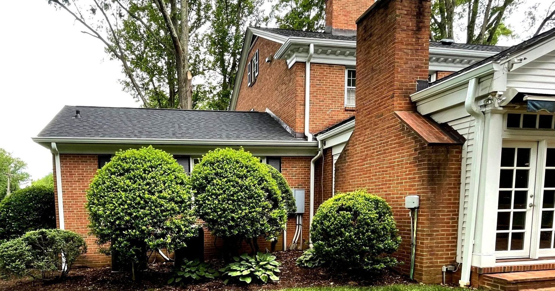 A large brick house with a chimney and a porch surrounded by bushes and trees.