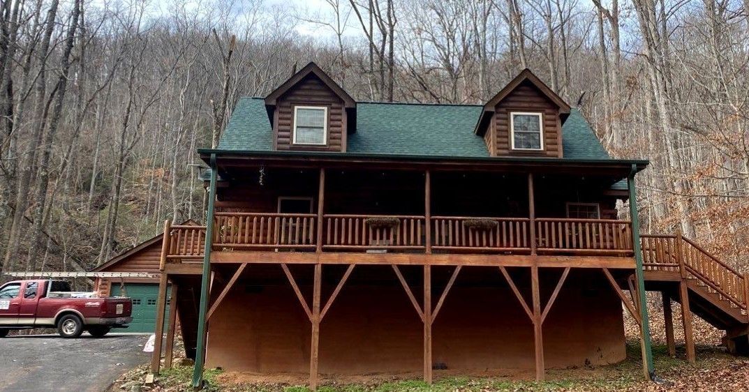 A large log cabin with a green roof and a large deck in the middle of the woods.