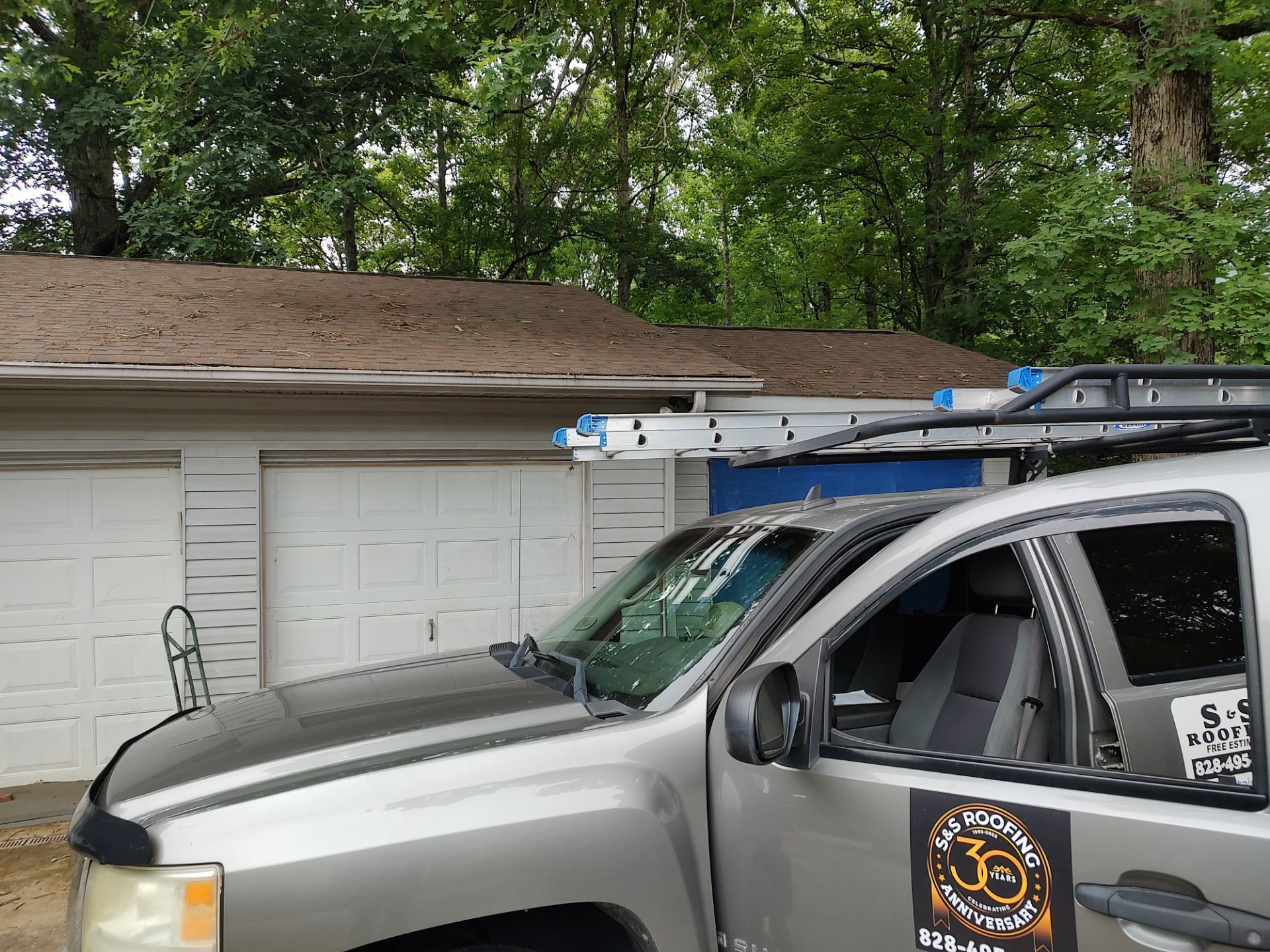 A silver truck with a ladder on top of it is parked in front of a garage.