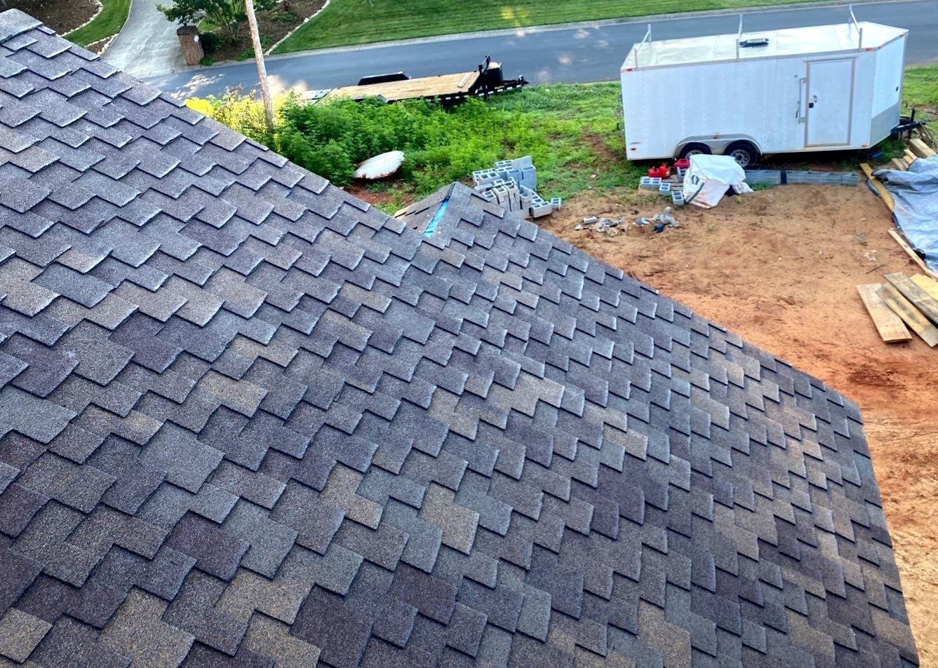 A roof with shingles being installed on a house with a trailer in the background.