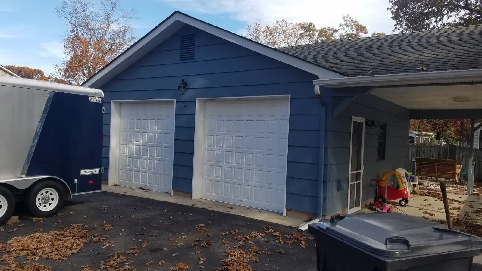 A blue garage with white garage doors and a trailer parked in front of it.