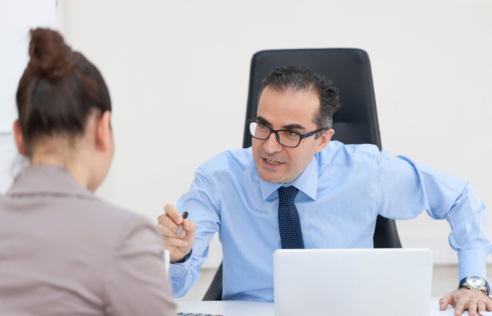 Man in blue shirt gestures, speaking to a person from the back. Inside office setting with laptop.