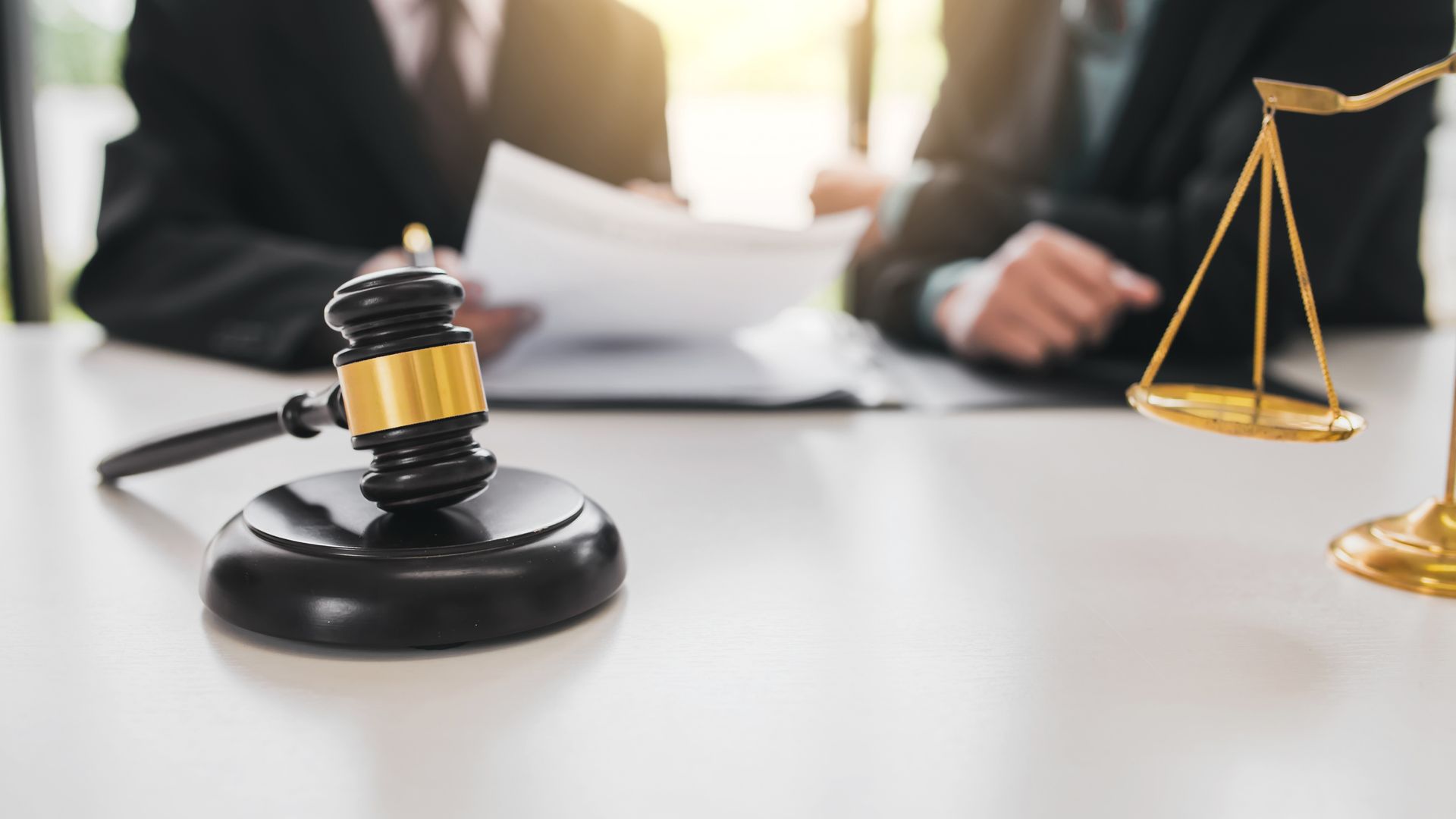 Gavel and balance scales on a table, with two people in suits reviewing documents in the background. Gavel and balance scales on a table, with two people in suits reviewing documents in the background.