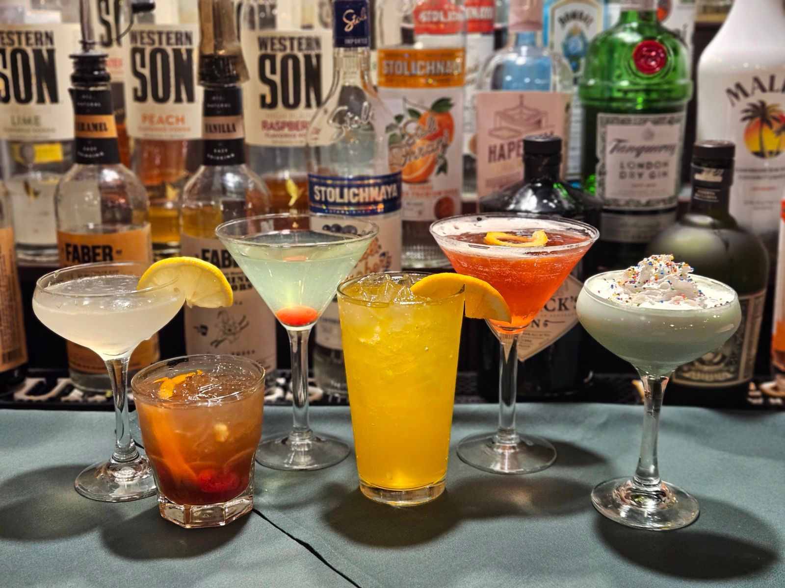 bar top with six specialty cocktails in front of bottles of alcohol
