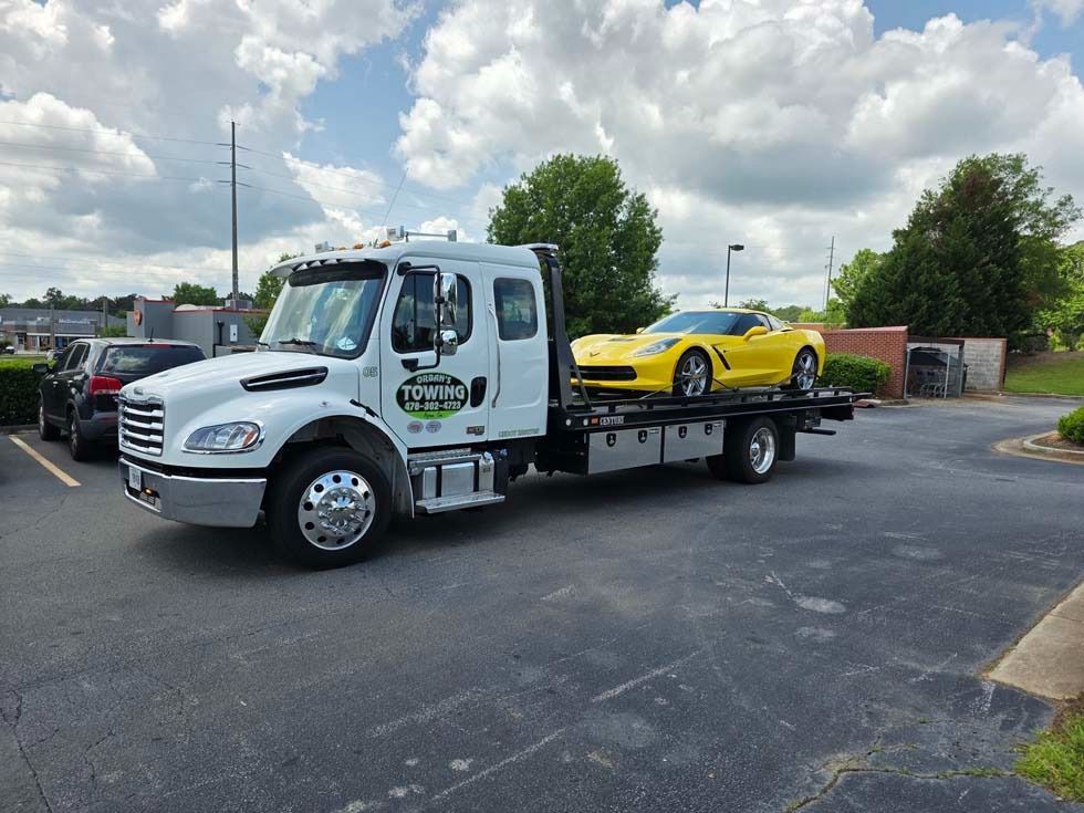 White tow truck carrying a yellow Corvette on a paved parking lot under a cloudy sky.