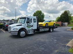 White tow truck carrying a yellow Corvette on a paved parking lot under a cloudy sky.