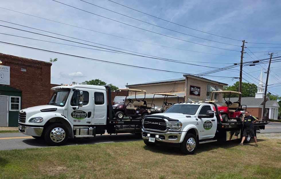 Two white tow trucks parked roadside; one with a car loaded, the other with a person near the back. Sunny day.