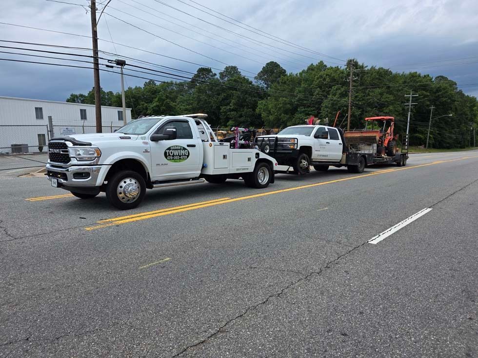 Tow truck towing a white pickup truck with an orange piece of machinery on a trailer. Roadside scene.