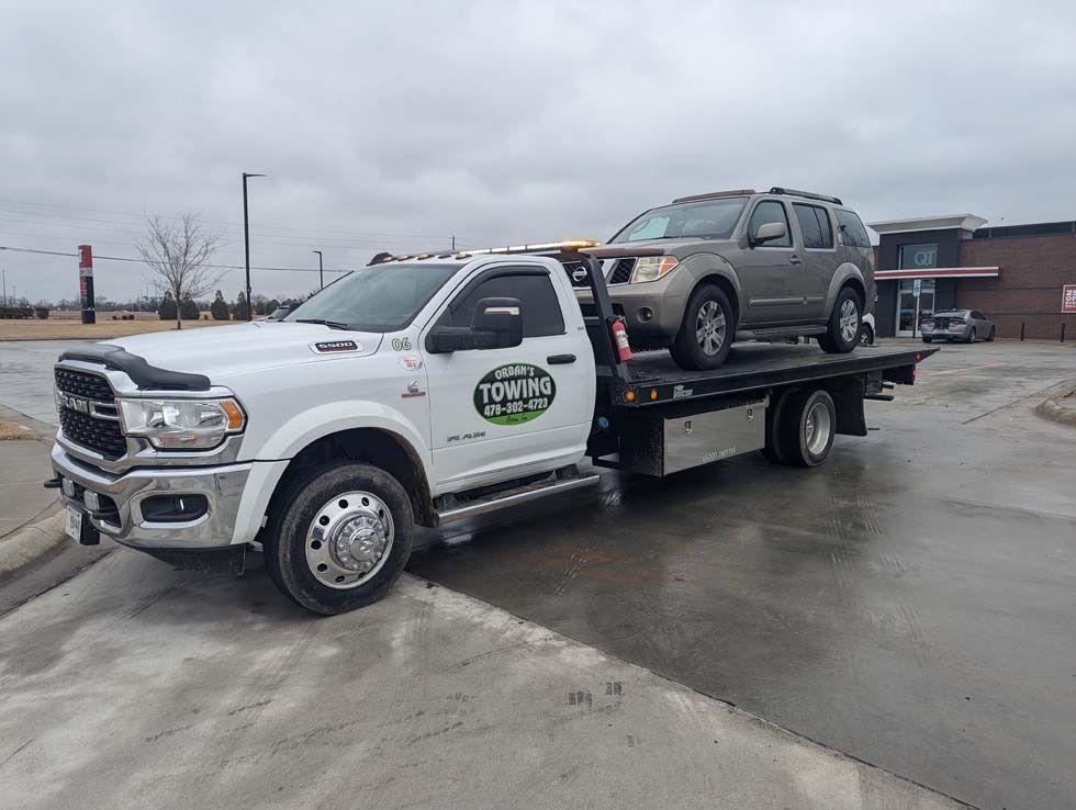 White tow truck carrying a gray SUV on a flatbed, parked near a building on a wet day.