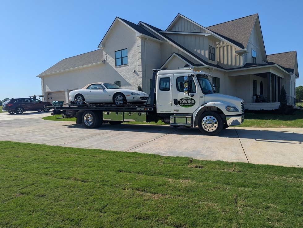 Tow truck with white car on flatbed in front of a large house on a sunny day.