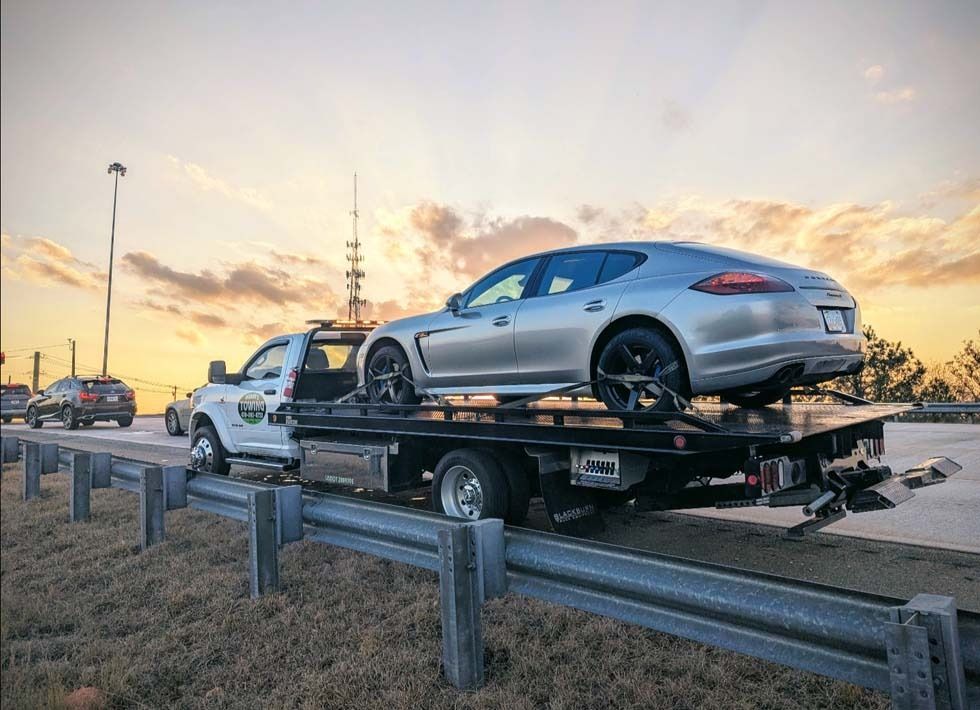 Silver Porsche sedan on a flatbed tow truck on a roadside, under an orange and yellow sunset.