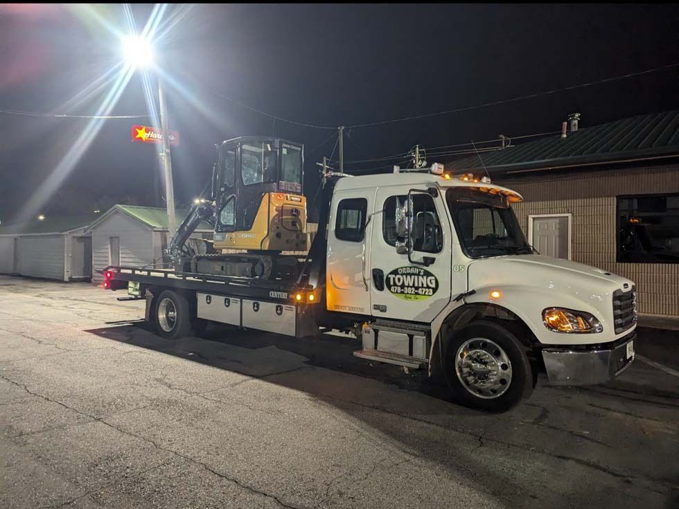Tow truck with a yellow excavator on its flatbed at night, parked near buildings.