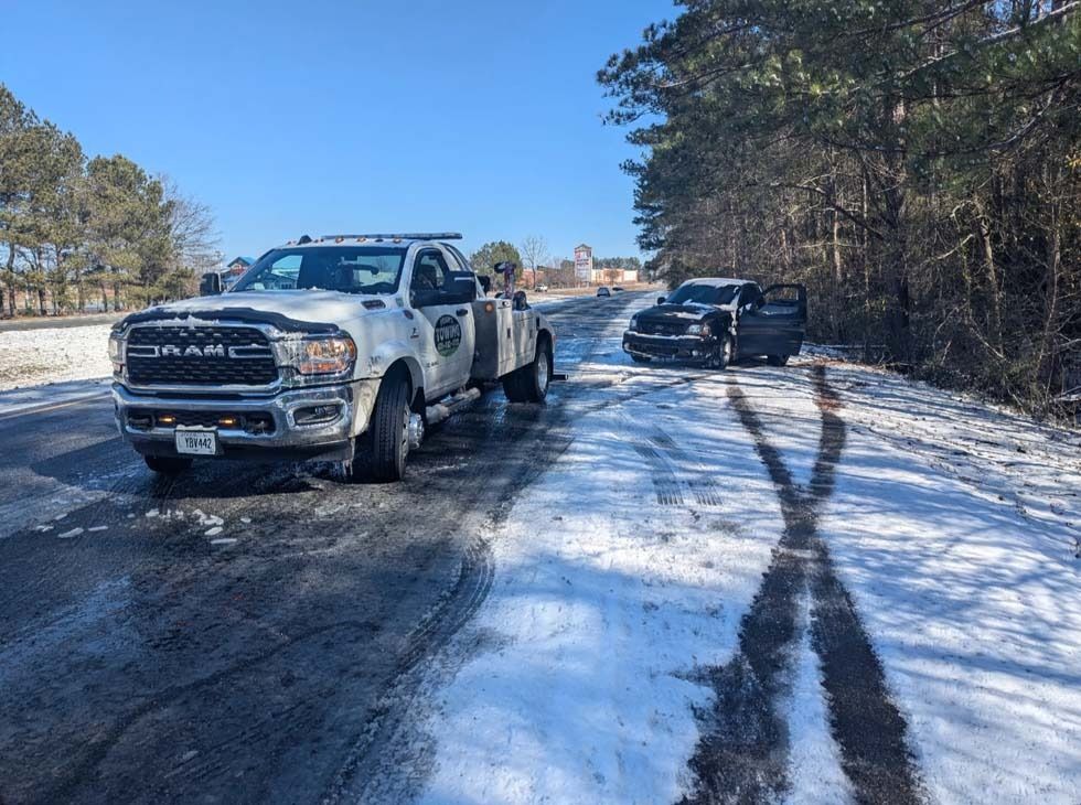 Tow truck at snowy roadside assisting a disabled black vehicle on a sunny day.