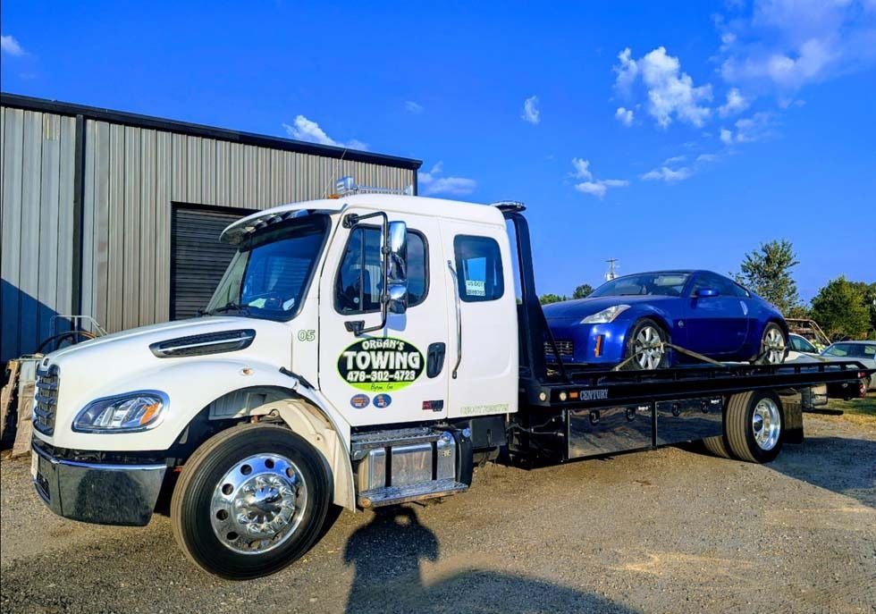White tow truck hauling a blue sports car outdoors under a partly cloudy sky.