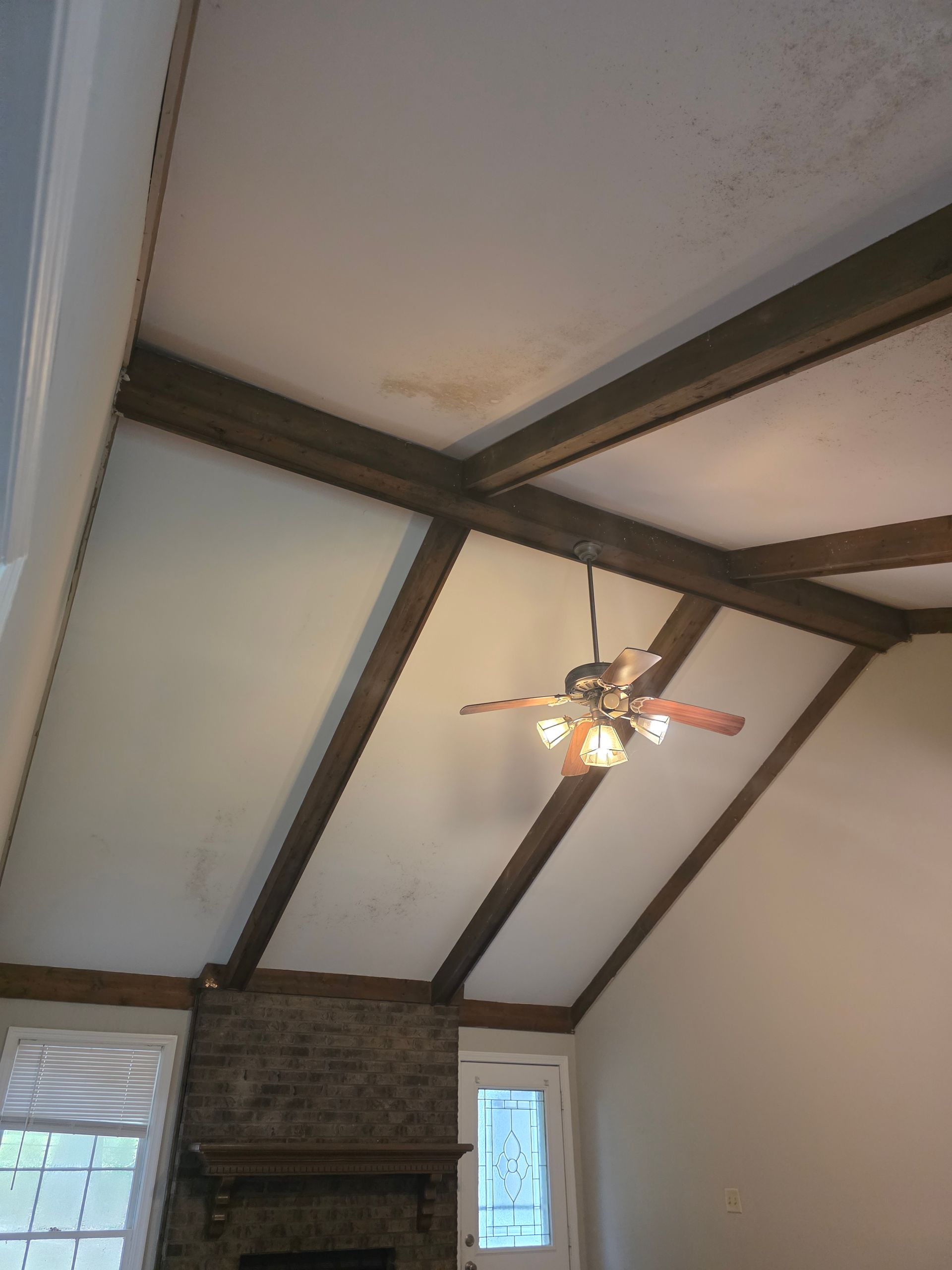 Angled white ceiling with dark beams, a ceiling fan, and a brick fireplace.