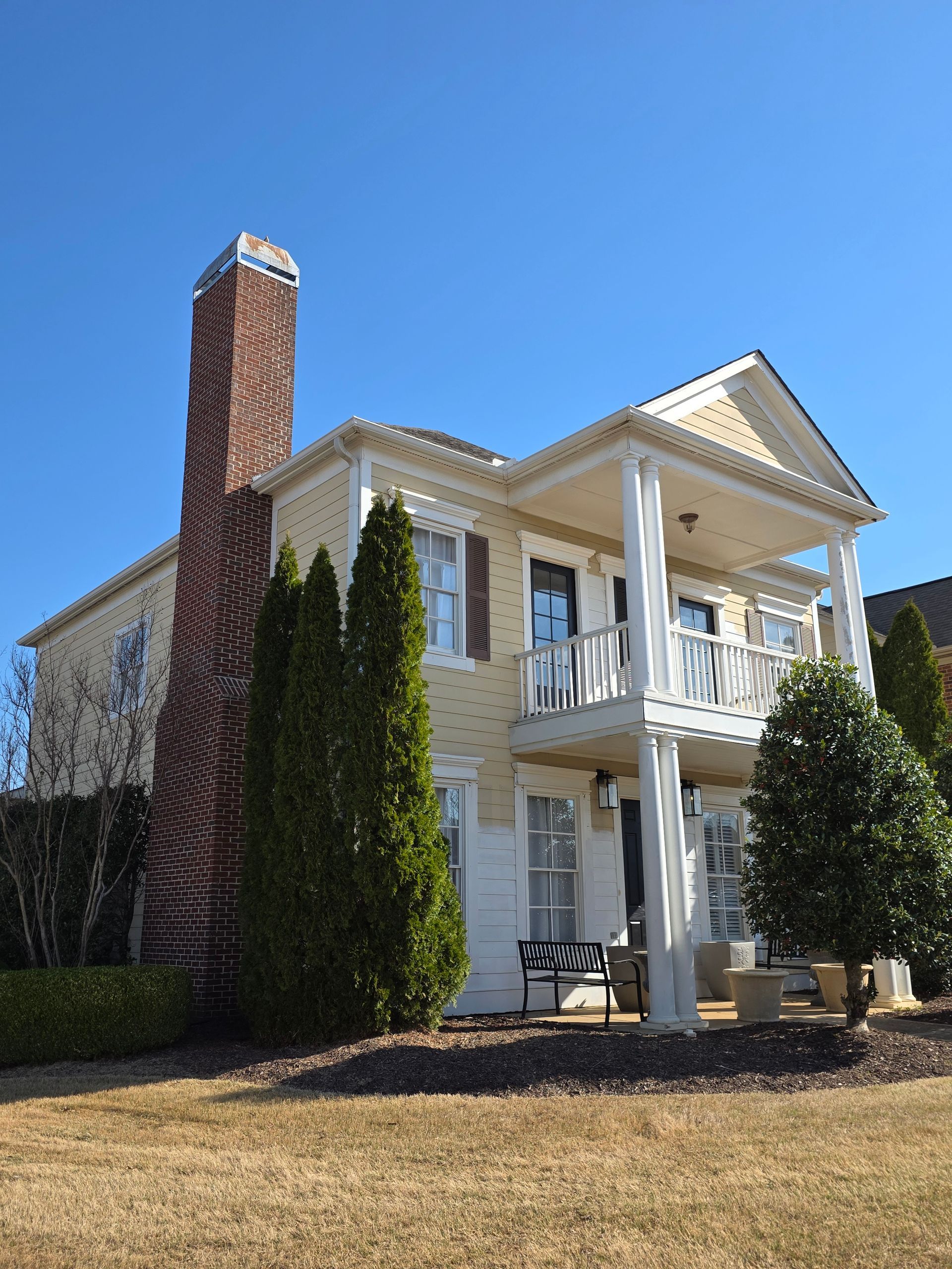 Two-story yellow house with white trim, a brick chimney, and a second-story porch under a blue sky.