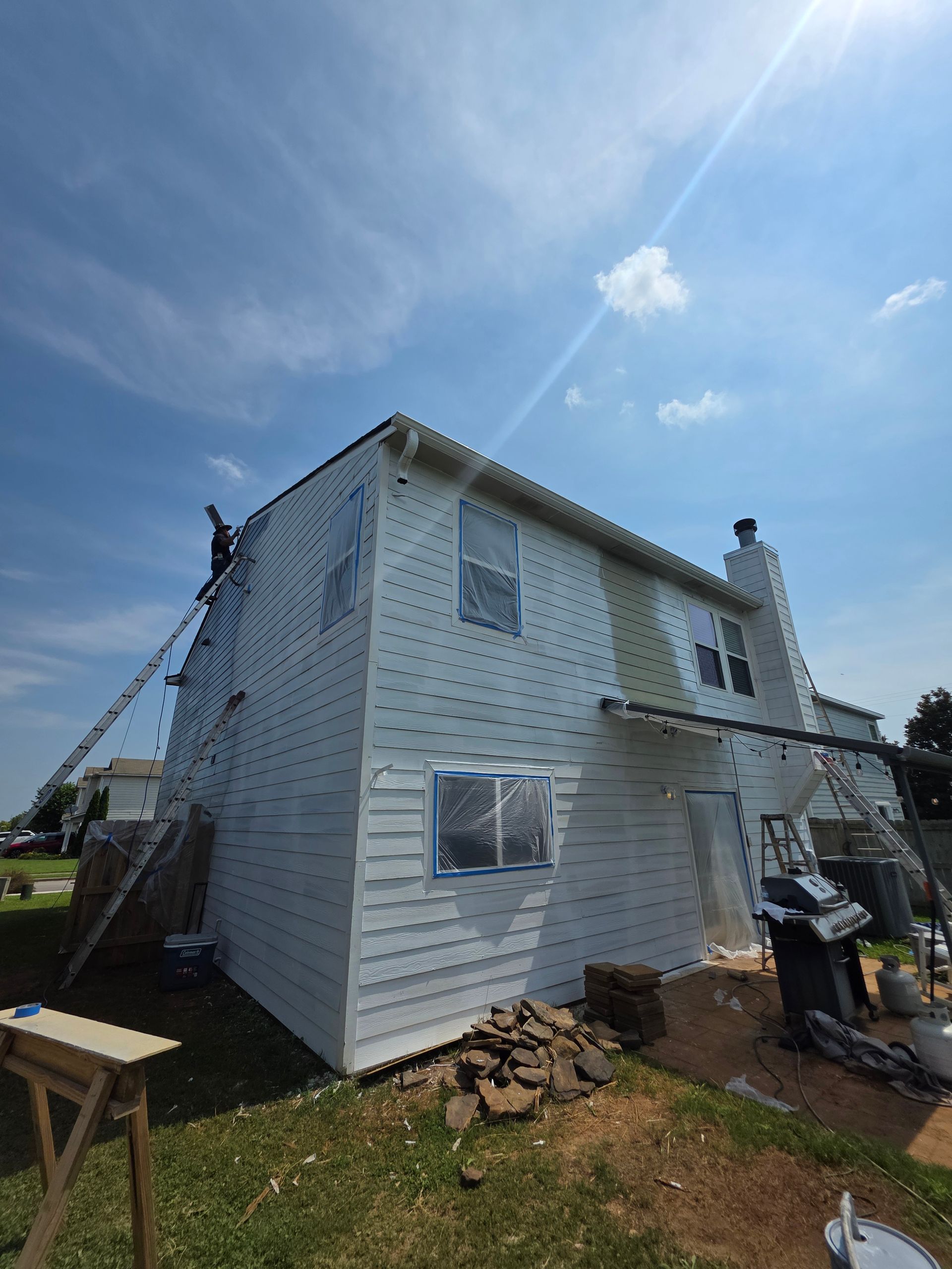 House with white siding being worked on under a sunny blue sky. A person on a ladder.